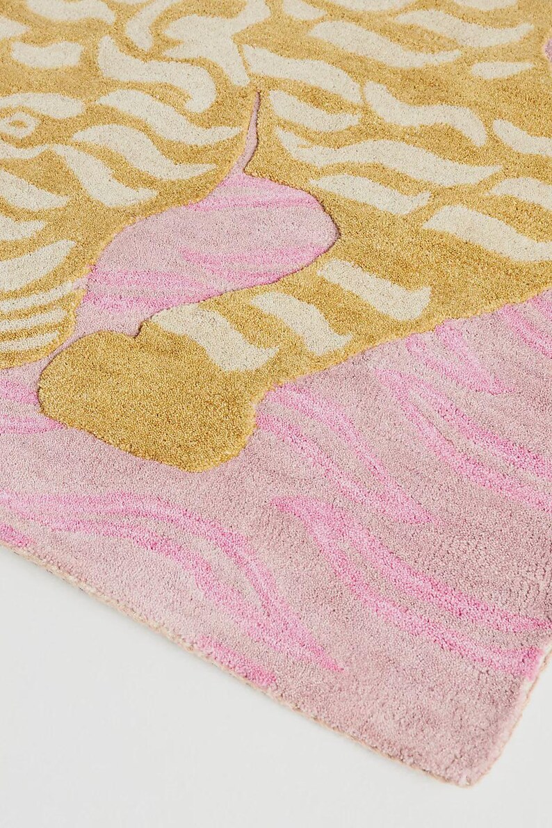 May include: A close-up of a rug with a textured surface. The rug features a design with gold and off-white wavy patterns on a pink background. The rug has a soft, plush appearance.