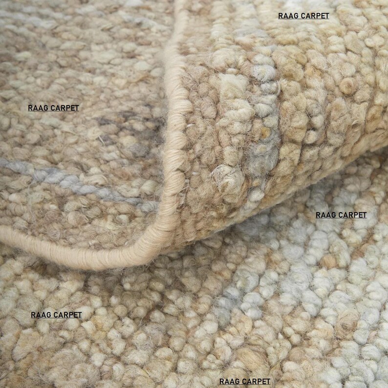 May include: Close-up of a textured, neutral-toned rug with a rolled edge. The rug has a nubby, woven appearance with variations in beige, cream, and gray. The text "RAAG CARPET" is visible in several places.