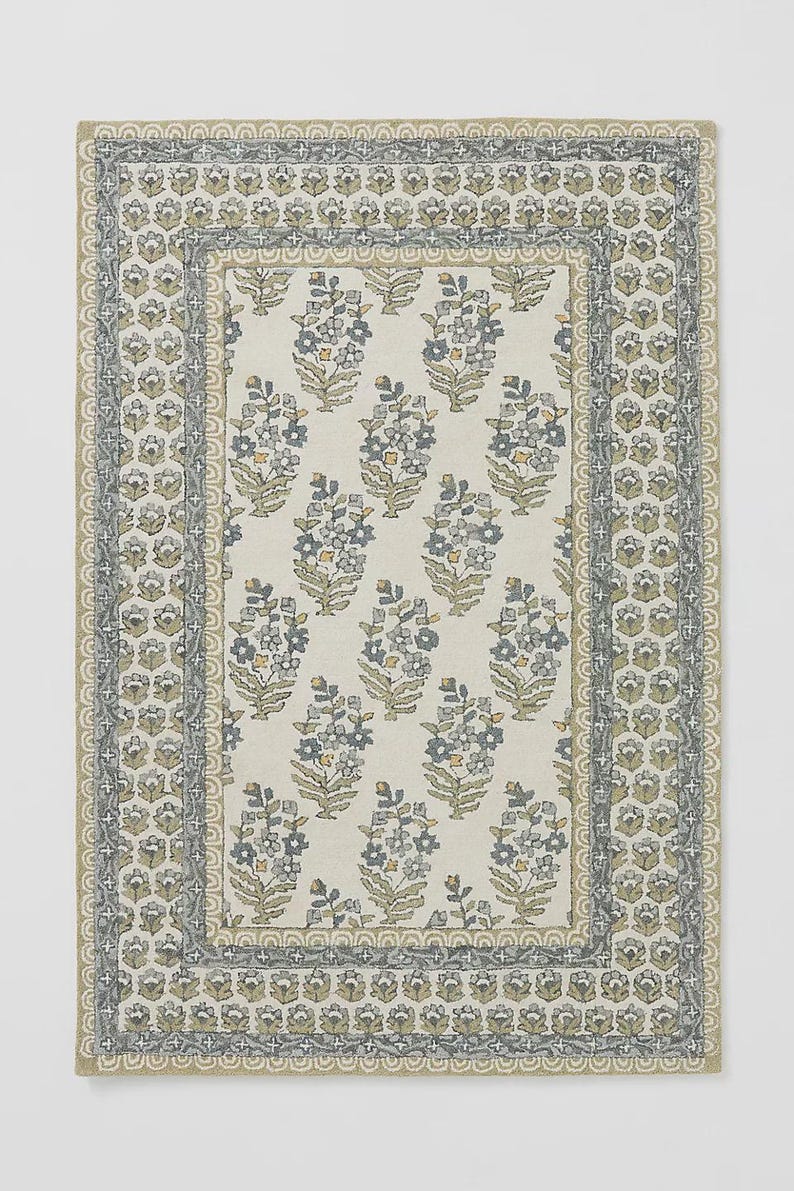 May include: Rectangular floral patterned rug with a cream background and a border. The rug features repeating clusters of blue and gray flowers with yellow accents. The border has a repeating floral design in shades of gray and green.