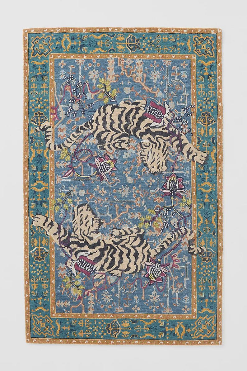 Puede incluir: Una alfombra rectangular con fondo azul y borde detallado. Dos tigres con rayas negras y detalles florales adornan la alfombra. La alfombra tiene un patr&oacute;n detallado y un borde marr&oacute;n.