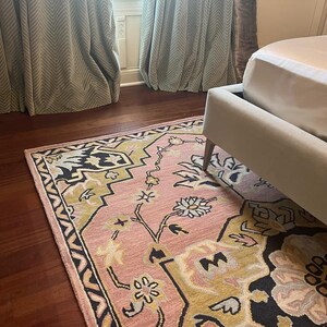 May include: A pink and gold area rug with a floral design and black border sits on a dark wood floor. The rug is partially under a bed frame. Pale green curtains are visible in the background.
