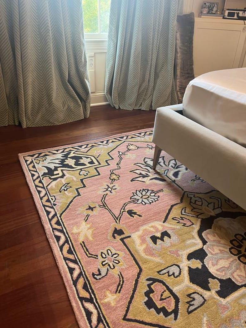 May include: A pink and gold area rug with a floral design and black border sits on a dark wood floor. The rug is partially under a bed frame. Pale green curtains are visible in the background.