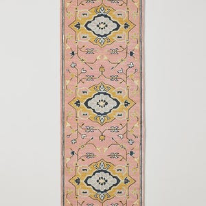 May include: A rectangular pink rug with an intricate geometric pattern. The design features repeating floral and geometric motifs in yellow, navy, and light blue. The rug is a runner style, ideal for hallways or entryways.