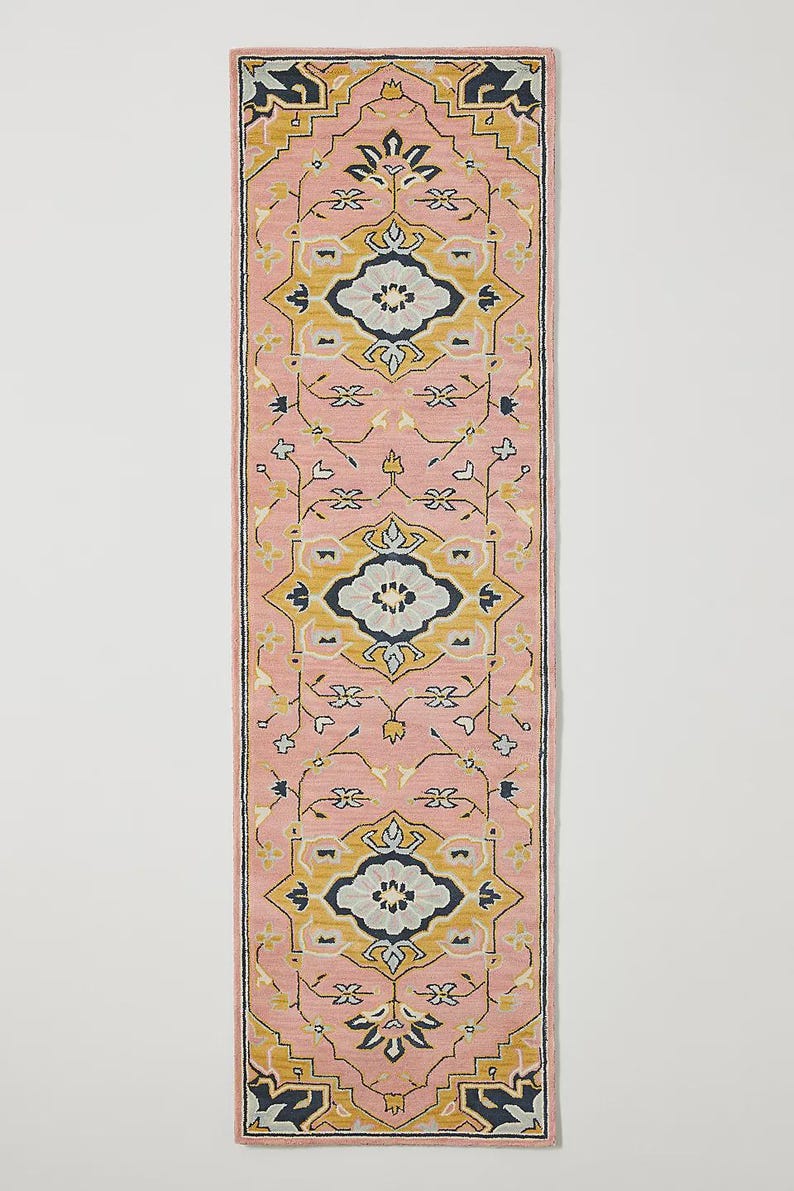 May include: A rectangular pink rug with an intricate geometric pattern. The design features repeating floral and geometric motifs in yellow, navy, and light blue. The rug is a runner style, ideal for hallways or entryways.