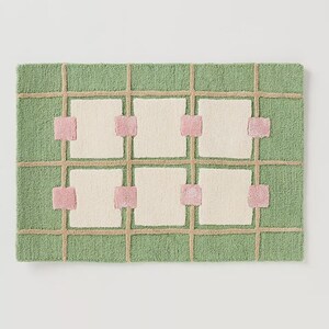 May include: A rectangular rug with a green background and a grid pattern. The grid is made of tan lines, with off-white squares and pink accents. The rug has a textured, woven appearance.