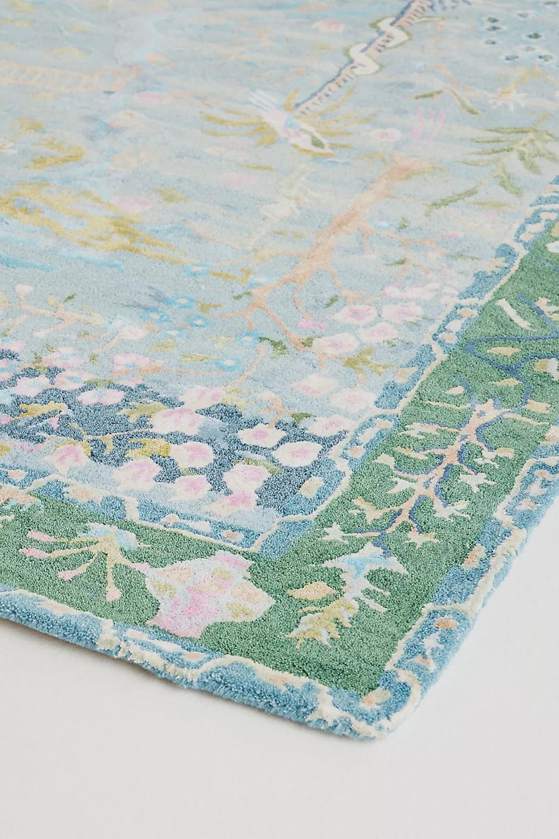 May include: A light blue rug with a floral design and a green border. The rug features a pattern of pink and yellow flowers, birds, and branches. The border is a darker green with a repeating leaf pattern.