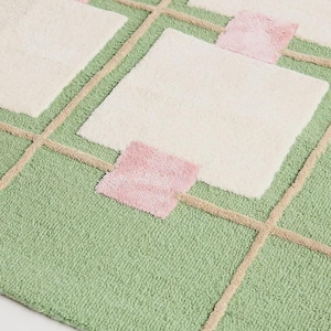 May include: A close-up of a geometric rug with a green background, cream-colored squares, and pink accents. The rug features a grid-like pattern with beige lines. The texture appears soft and plush.