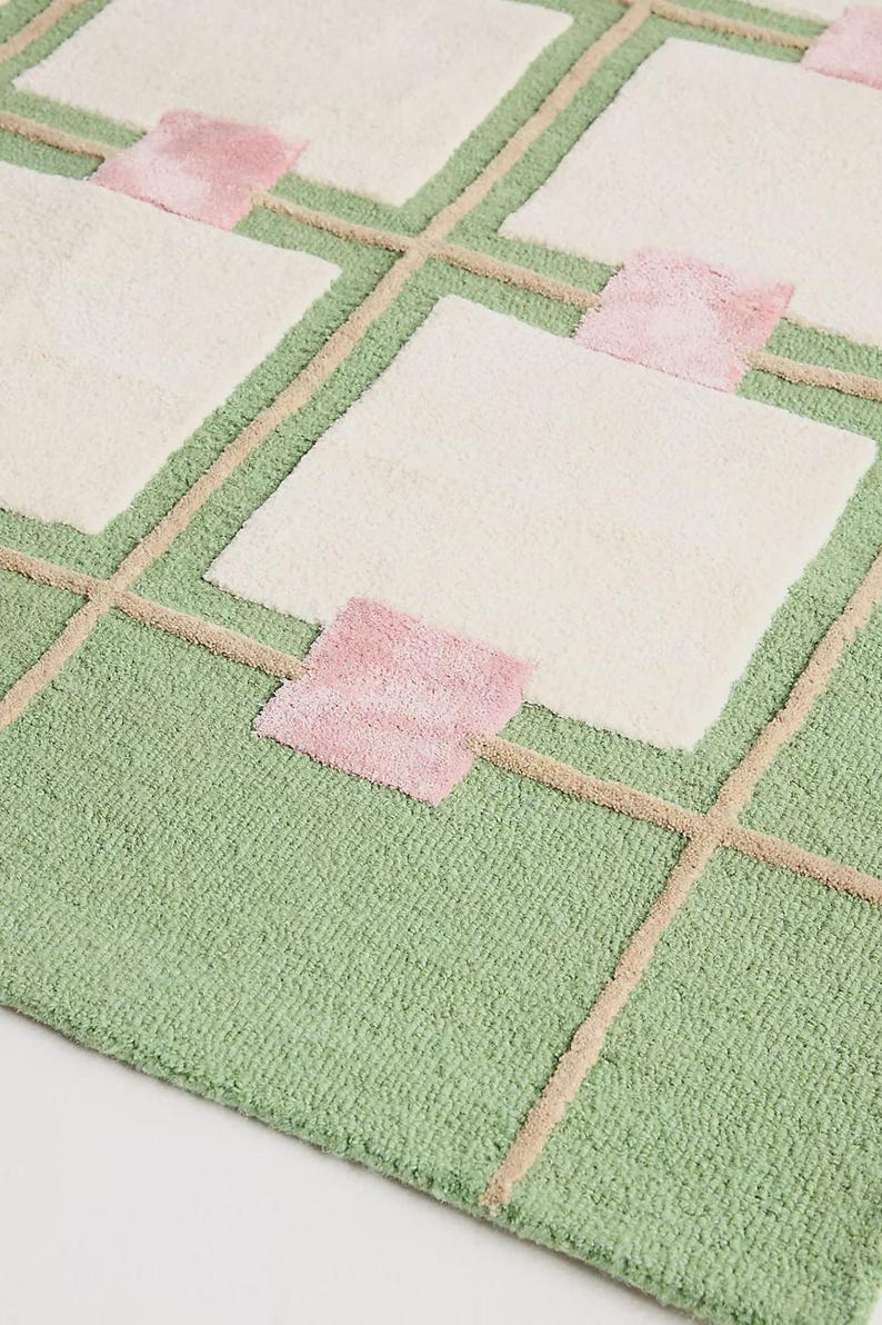 May include: A close-up of a geometric rug with a green background, cream-colored squares, and pink accents. The rug features a grid-like pattern with beige lines. The texture appears soft and plush.