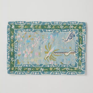 May include: Rectangular rug with a light blue center featuring floral and bird designs in pink, green, and white. The rug has a green border with a repeating floral pattern. The rug is approximately 36 inches long and 24 inches wide.