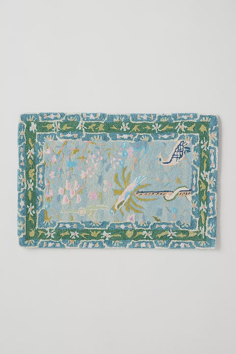 May include: Rectangular rug with a light blue center featuring floral and bird designs in pink, green, and white. The rug has a green border with a repeating floral pattern. The rug is approximately 36 inches long and 24 inches wide.