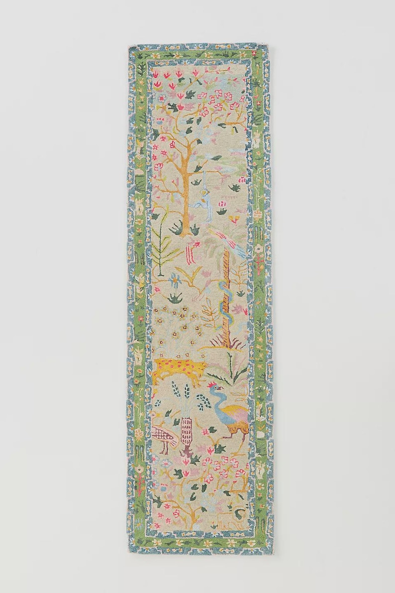 May include: A rectangular, decorative runner featuring a whimsical, colorful design. The runner showcases a light beige background with a detailed pattern of trees, flowers, birds, and a leopard. The edges are bordered with a green and blue floral pattern.