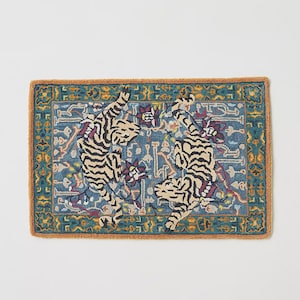 Puede incluir: Alfombra rectangular con fondo azul y borde marr&oacute;n. La alfombra presenta dos tigres estilizados con rayas negras y beige, rodeados de patrones florales y geom&eacute;tricos. Un elemento decorativo para el hogar.