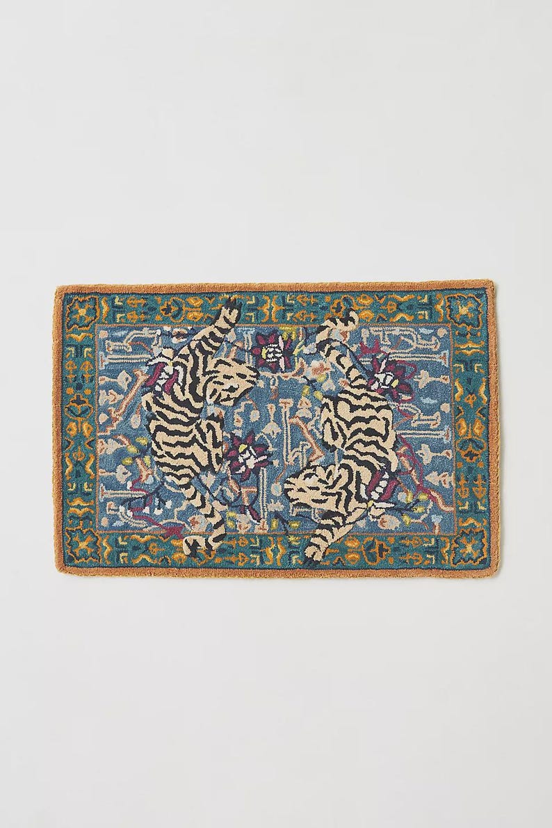 Puede incluir: Alfombra rectangular con fondo azul y borde marr&oacute;n. La alfombra presenta dos tigres estilizados con rayas negras y beige, rodeados de patrones florales y geom&eacute;tricos. Un elemento decorativo para el hogar.