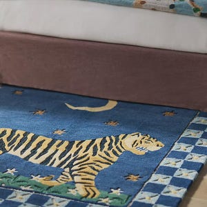 Op de afbeelding: Een blauw tapijt met een tijgerontwerp. De tijger is geel en zwart, tegen een blauwe achtergrond met sterren en een halve maan. Het tapijt heeft een geblokte rand en ligt voor een bed.