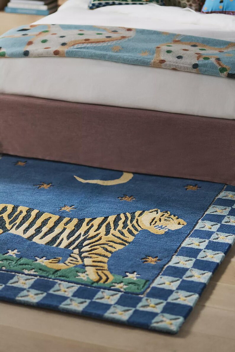 Op de afbeelding: Een blauw tapijt met een tijgerontwerp. De tijger is geel en zwart, tegen een blauwe achtergrond met sterren en een halve maan. Het tapijt heeft een geblokte rand en ligt voor een bed.
