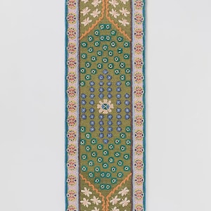 May include: A rectangular, embroidered runner with a green central design. The runner features a repeating floral pattern in white, orange, and blue, with a blue border. The central design includes a diamond shape with floral and geometric elements.