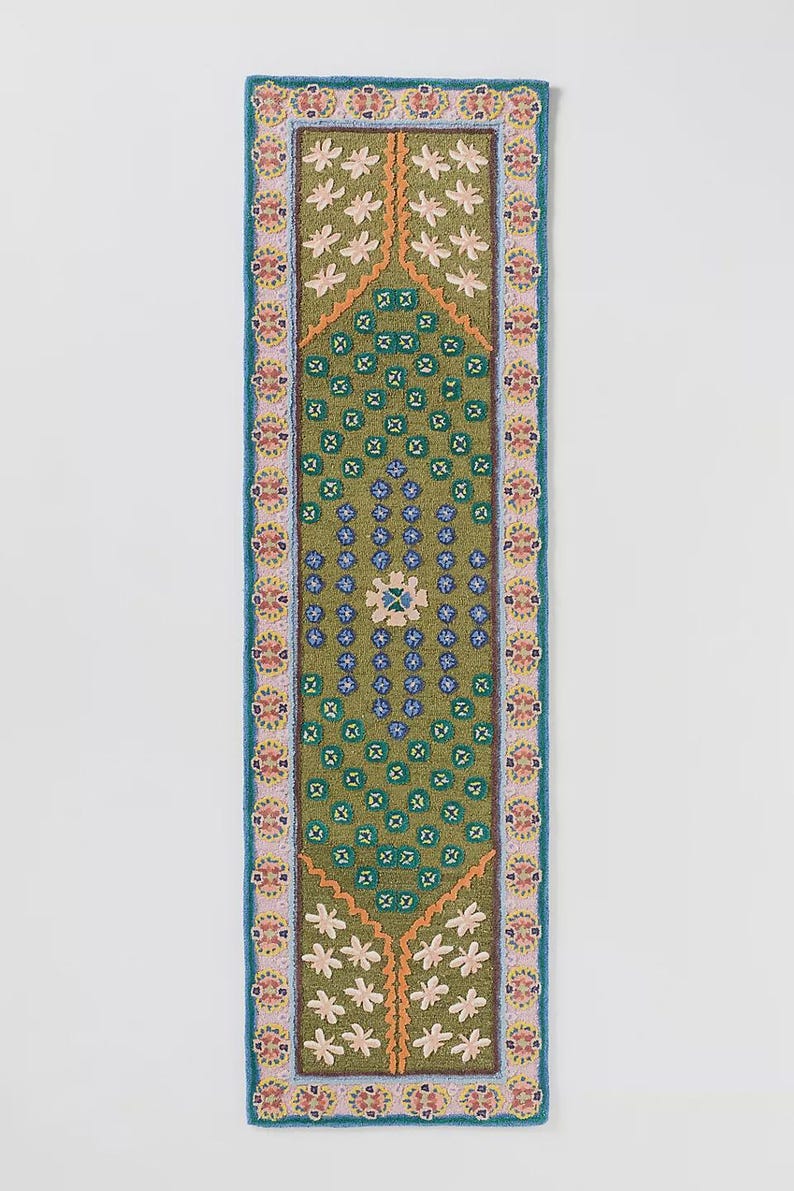 May include: A rectangular, embroidered runner with a green central design. The runner features a repeating floral pattern in white, orange, and blue, with a blue border. The central design includes a diamond shape with floral and geometric elements.