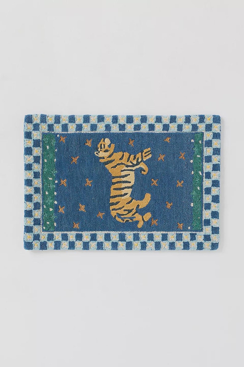 Op de afbeelding: Blauw rechthoekig tapijt met een tijgermotief. De tijger is geel en zwart, tegen een blauwe achtergrond met gouden sterren. Het tapijt heeft een blauw-witte geblokte rand.