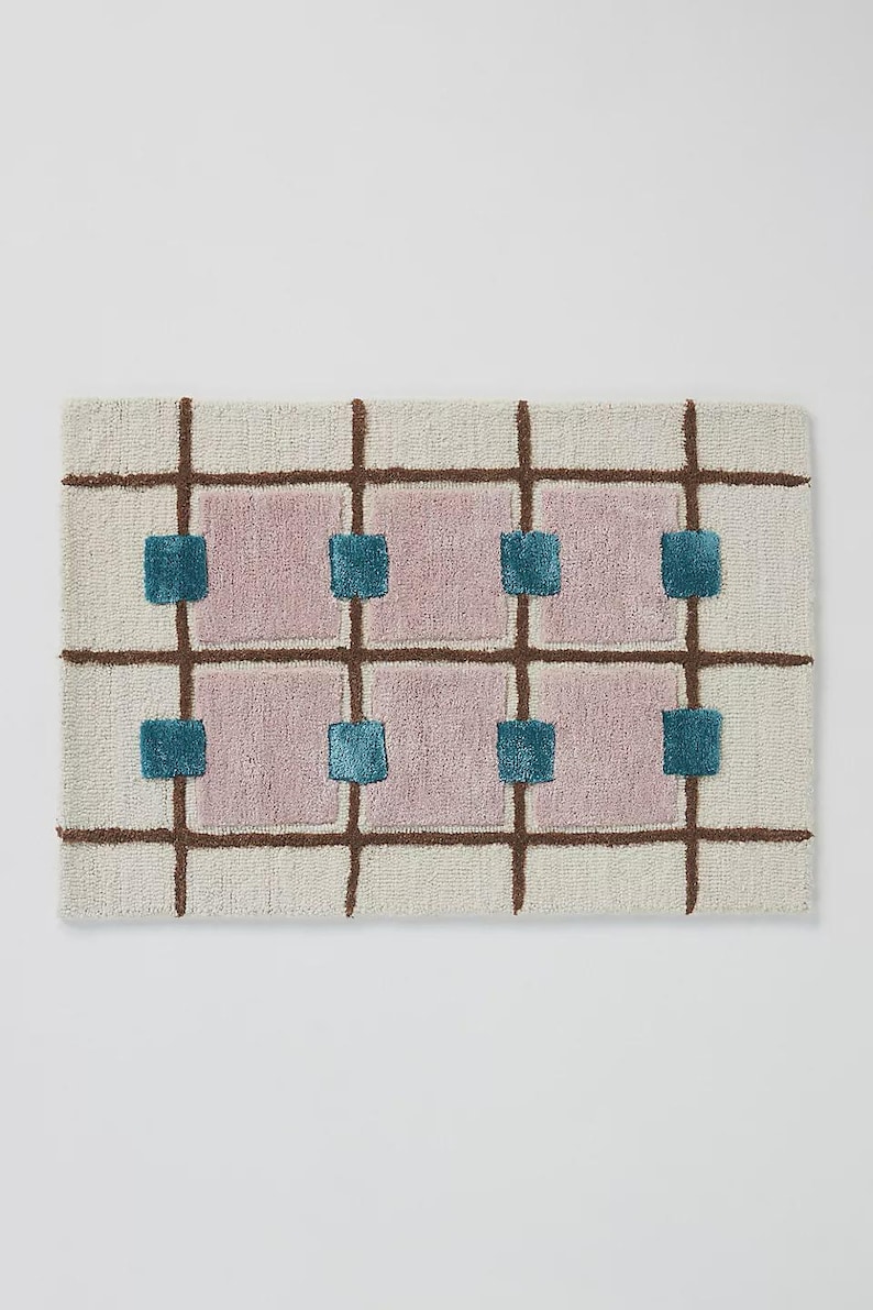 Puede incluir: Alfombra rectangular con fondo crema, l&iacute;neas de cuadr&iacute;cula marrones y cuadrados rosas. Cuadrados turquesa se colocan dentro de los cuadrados rosas. La alfombra tiene un aspecto texturizado y tejido, adecuada para una sala de estar o dormitorio.
