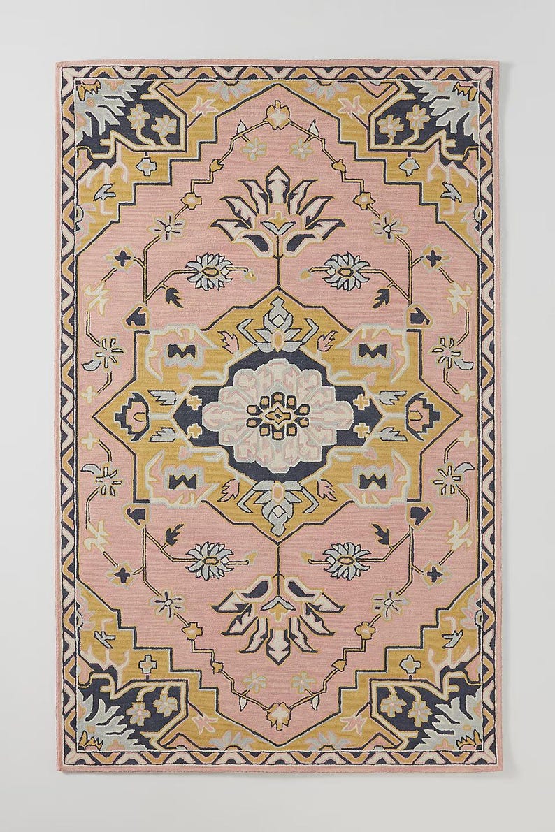 May include: A rectangular rug with a pink background and a detailed geometric pattern. The design incorporates yellow, navy, and white floral and geometric shapes. The rug has a border with a repeating pattern.