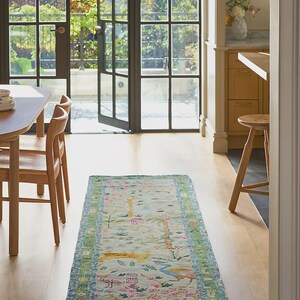 May include: A rectangular, light-colored rug with a detailed floral and bird pattern. The rug has a green and blue border, and is placed on a hardwood floor. The room has large windows and wooden chairs.