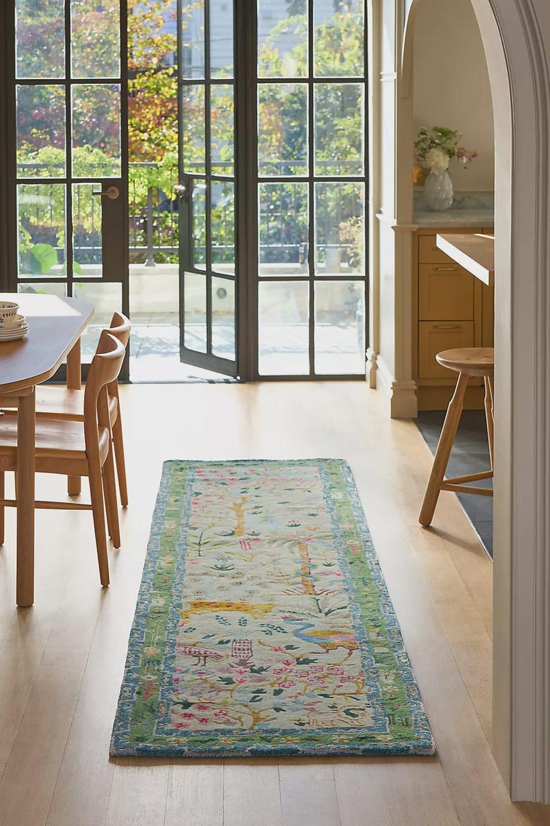 May include: A rectangular, light-colored rug with a detailed floral and bird pattern. The rug has a green and blue border, and is placed on a hardwood floor. The room has large windows and wooden chairs.