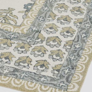 May include: A rectangular rug with a floral pattern in shades of cream, gray, and gold. The rug features a detailed border with a repeating floral motif and a scalloped edge. The design is symmetrical and creates a classic, elegant look.