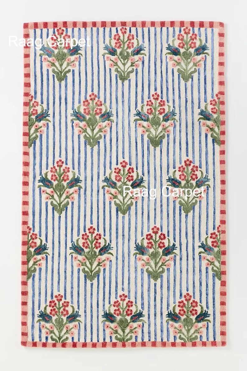 May include: Rectangular rug with a blue and white striped background. The rug features a repeating floral pattern in pink, green, and blue. The border is a red and pink striped pattern. The text "Raag Carpet" is visible.