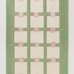 May include: A rectangular rug with a green background and a grid pattern. The grid is made of tan lines, with off-white squares and pink rectangular accents. The rug has a soft, textured appearance, suitable for home decor.
