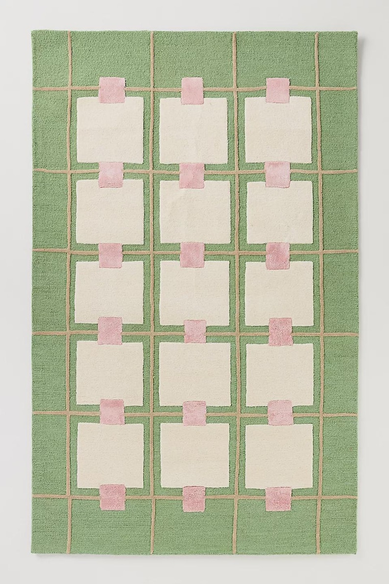 May include: A rectangular rug with a green background and a grid pattern. The grid is made of tan lines, with off-white squares and pink rectangular accents. The rug has a soft, textured appearance, suitable for home decor.