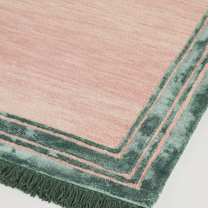 May include: A rectangular rug with a pink center and a teal green border. The border has a layered design with a fringe along one edge. The rug's texture appears soft and plush.