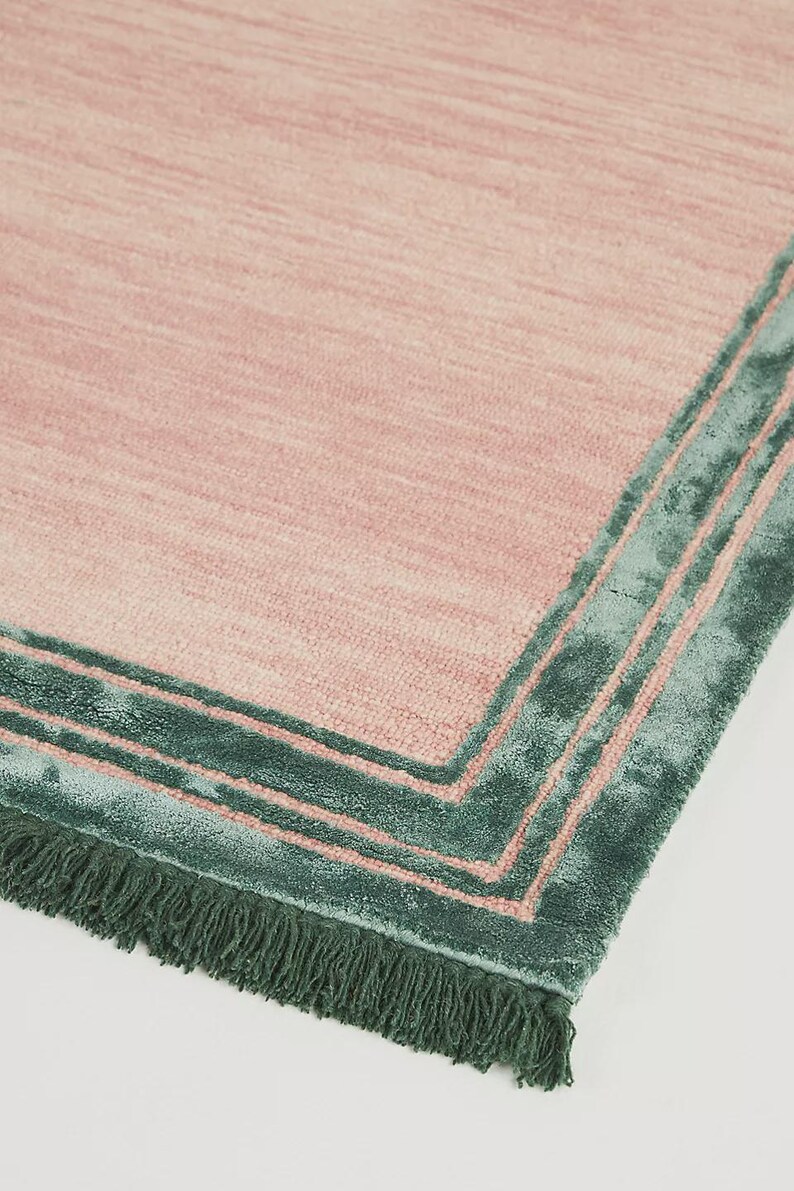 May include: A rectangular rug with a pink center and a teal green border. The border has a layered design with a fringe along one edge. The rug's texture appears soft and plush.