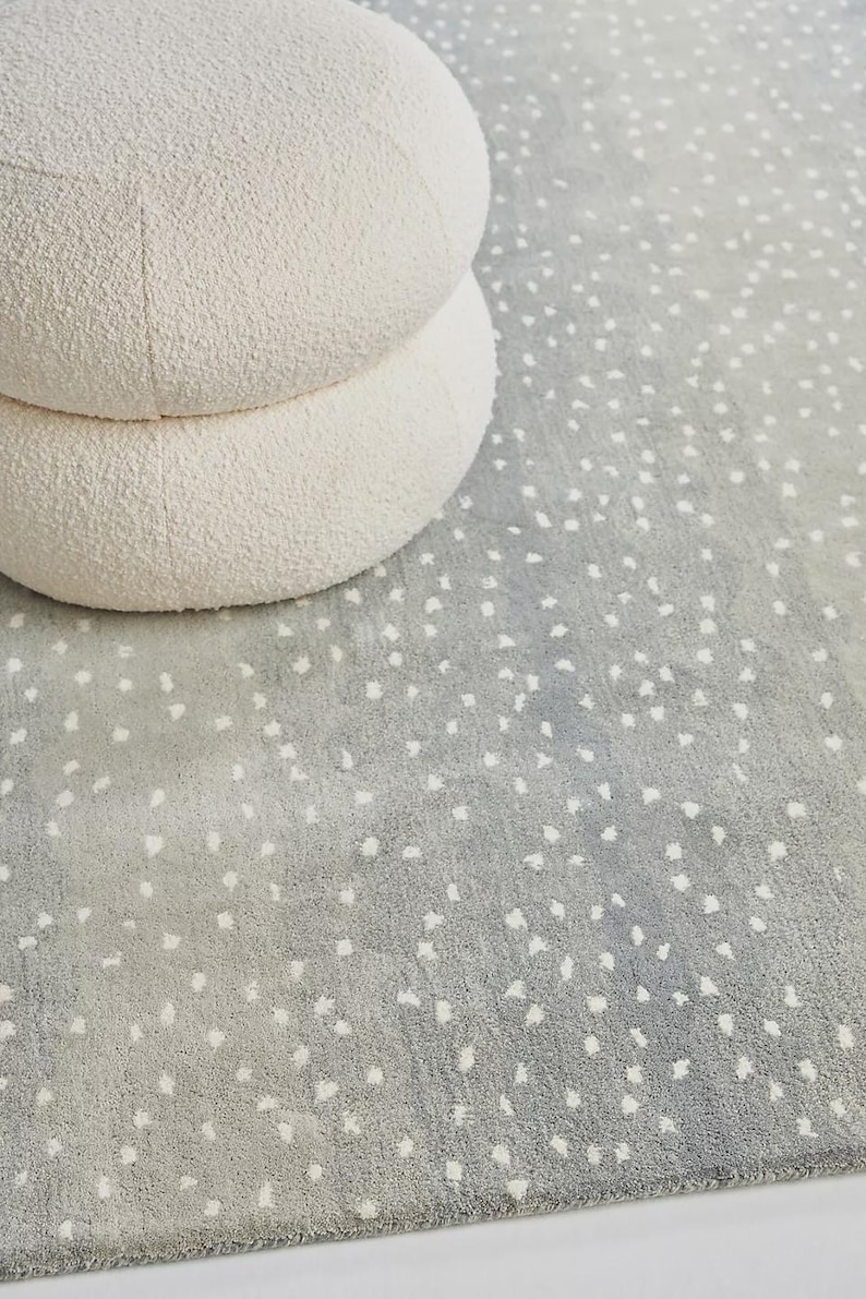 May include: A light gray rug with a speckled pattern and a white, round, two-tiered ottoman. The rug has a soft, textured appearance, and the ottoman is made of a plush, cream-colored material. The overall aesthetic is modern and minimalist.