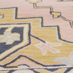 May include: Close-up of a pink rug with a geometric pattern. The design features shapes in shades of yellow, navy blue, and light pink. The rug has a soft, textured appearance, suggesting a comfortable and decorative home item.