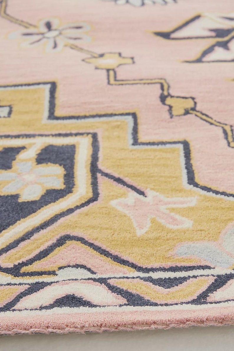 May include: Close-up of a pink rug with a geometric pattern. The design features shapes in shades of yellow, navy blue, and light pink. The rug has a soft, textured appearance, suggesting a comfortable and decorative home item.