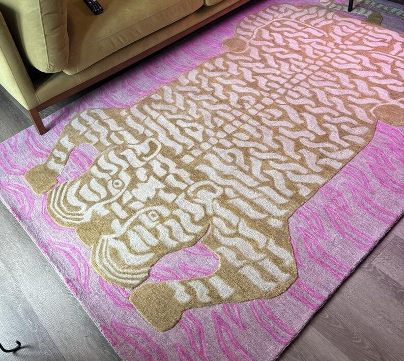 May include: A rectangular rug with a tiger design. The rug features a tan tiger shape with a white pattern on a pink background with pink wave-like patterns. The rug is placed on a wooden floor near a beige sofa.