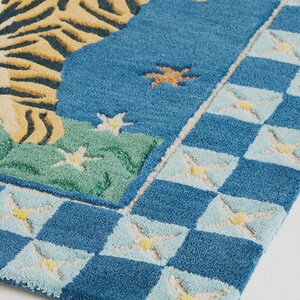 Op de afbeelding: Een blauw tapijt met een tijgerontwerp. Het tapijt heeft de kop en het lichaam van een tijger in gele en zwarte strepen tegen een blauwe achtergrond. De rand heeft een dambordpatroon met lichtblauwe en gele accenten. Het tapijt is een decoratief woonartikel.
