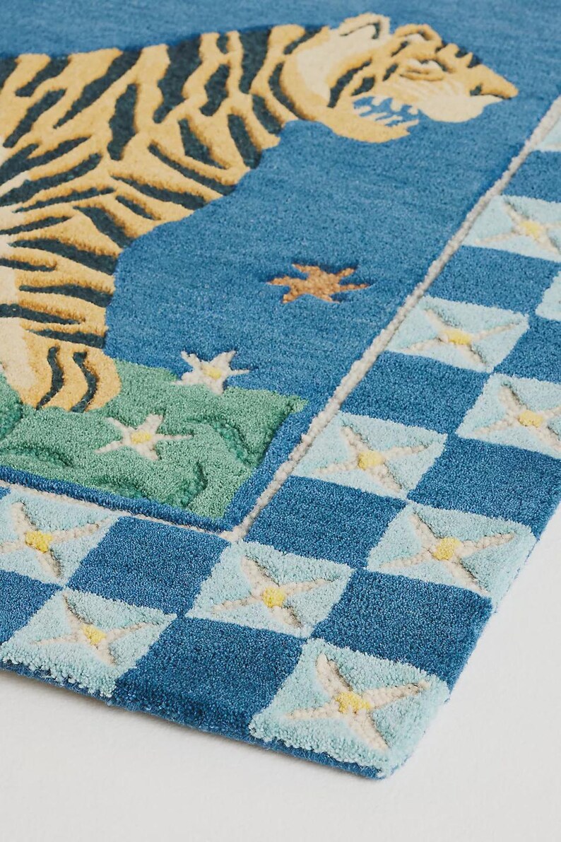 Op de afbeelding: Een blauw tapijt met een tijgerontwerp. Het tapijt heeft de kop en het lichaam van een tijger in gele en zwarte strepen tegen een blauwe achtergrond. De rand heeft een dambordpatroon met lichtblauwe en gele accenten. Het tapijt is een decoratief woonartikel.