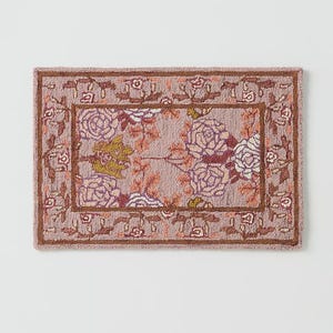 May include: Rectangular rug with a floral pattern. The rug features a light purple background with a central design of large roses in shades of purple, white, and yellow. A brown border frames the floral design, and the edges have a repeating floral motif.