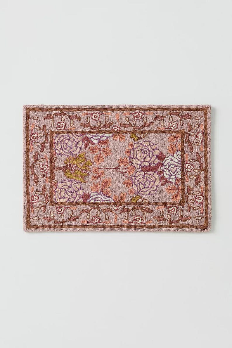 May include: Rectangular rug with a floral pattern. The rug features a light purple background with a central design of large roses in shades of purple, white, and yellow. A brown border frames the floral design, and the edges have a repeating floral motif.