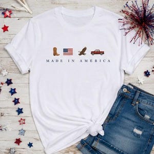 May include: A white short-sleeved t-shirt featuring a graphic print with a brown cowboy boot, an American flag, a bald eagle, and a red pickup truck. Below the icons, the text reads MADE IN AMERICA in a dark blue, sans-serif font.