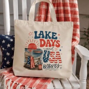 Può includere: Una borsa tote in tela color crema con una stampa grafica recante il testo "LAKE DAYS and USA WAVES". Il design include un sole rosso, una barca sull'acqua, una sedia in legno con un asciugamano a fantasia di bandiera e stelle rosse, bianche e blu.