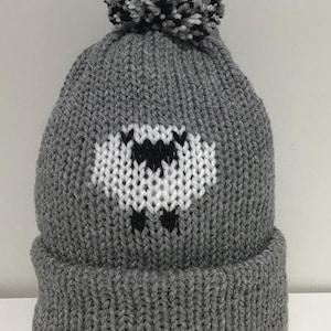 May include: A grey knitted beanie hat featuring a sheep motif in white and black. The hat has a folded brim and a pom-pom on top, ideal for keeping warm. The pom-pom is made of mixed black, white, and grey yarn.