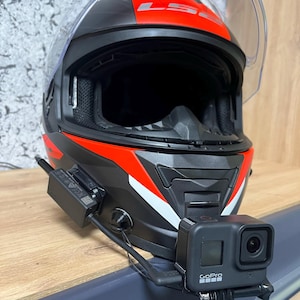 以下が含まれることがあります： 赤と白のストライプのデザインとクリアなシールドが付いた黒いオートバイのヘルメット。 GoProカメラが黒いマウントでヘルメットに取り付けられています。