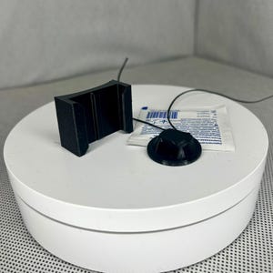 Puede incluir: Componentes negros impresos en 3D, incluyendo un soporte y una pieza circular con un cable, se muestran sobre una superficie blanca. También hay un pequeño paquete sellado. Parecen ser accesorios o piezas.