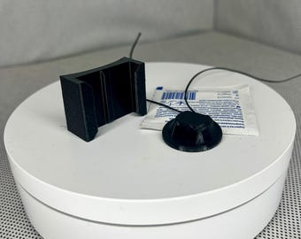 May include: Black 3D-printed components, including a bracket and a circular piece with a wire, are displayed on a white surface. A small, sealed packet is also present. The items appear to be accessories or parts.