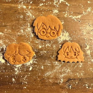 Könnte beinhalten: Drei braune Lebkuchen in Form von Comic-Gesichtern. Die Kekse liegen auf einer mit weißem Mehl bedeckten Holzoberfläche.