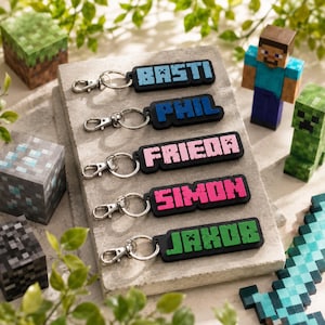 Könnte beinhalten: Fünf rechteckige Schlüsselanhänger mit Metallkarabinerhaken, jeweils mit schwarzer Basis und pixeliger Schrift in verschiedenen Farben. Die Namen BASTI, PHIL, FRIEDA, SIMON und JAKOB sind in einer blockartigen Schriftart dargestellt.