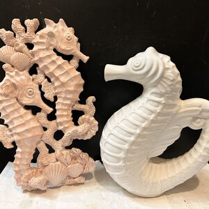 May include: Two ceramic seahorse sculptures, one white and one pink, with intricate details. The pink seahorse is part of a wall decor piece with coral and seashells.