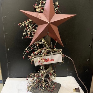 Pode incluir: Uma árvore de mesa rústica de madeira em forma de estrela com uma estrela de metal vermelha no topo. A árvore é decorada com ramos de bagas brancas e vermelhas e uma placa que diz "Phillies".